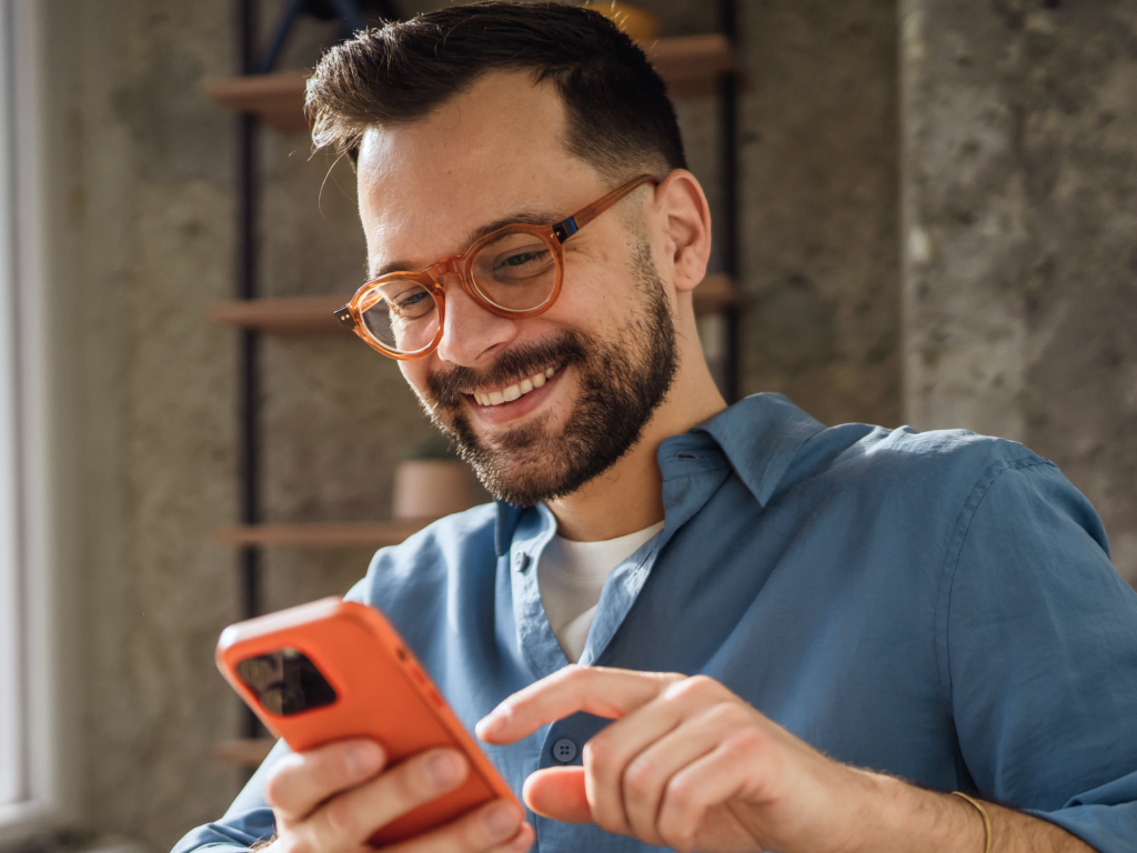 Uomo sorridente con occhiali arancioni e camicia azzurra usa lo smartphone, con una mensola sullo sfondo
