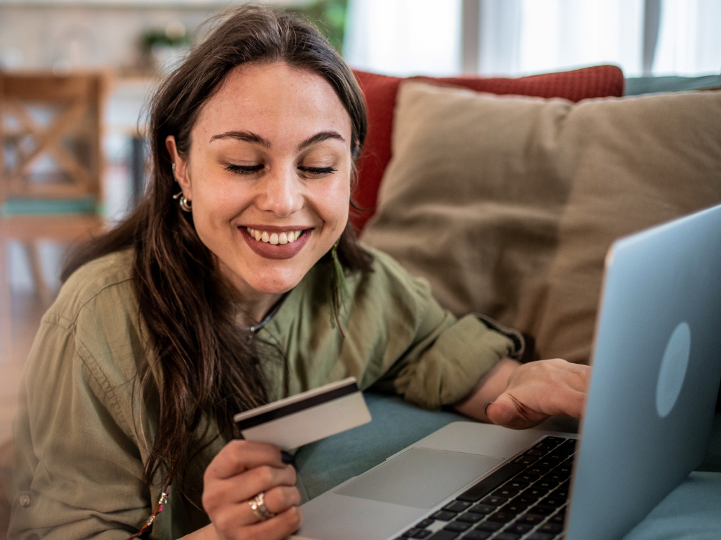 Giovane donna sorridente con una carta in mano fa acquisti online dal laptop, sdraiata sul divano di casa