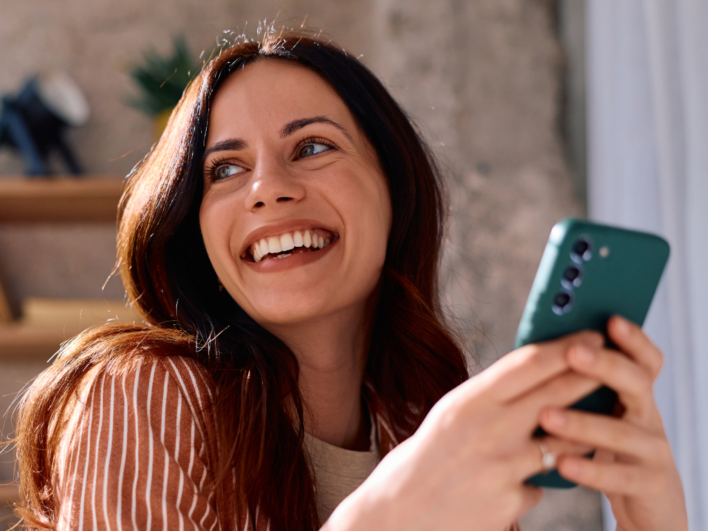 Giovane donna sorridente con lo smartphone in mano guarda di lato in una stanza luminosa
