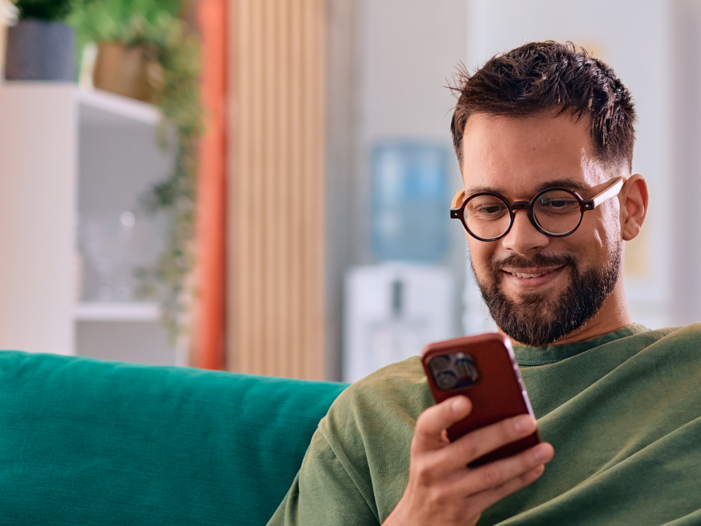 Uomo sorridente con occhiali tondi e maglione verde guarda lo smartphone seduto su un divano