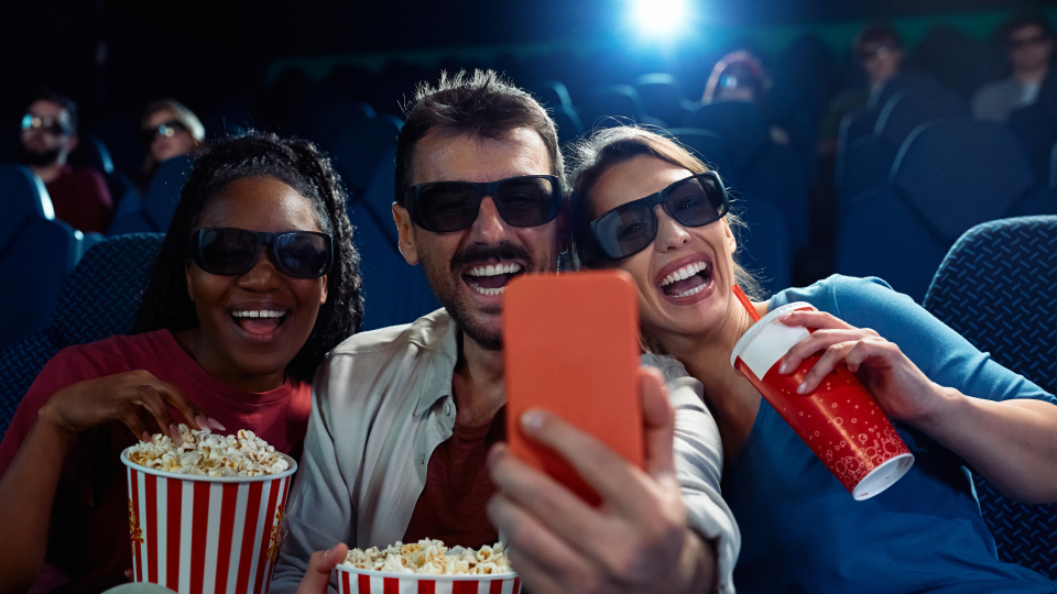 Tre amici con occhiali 3D ridono scattandosi un selfie al cinema, con popcorn e bibita in mano