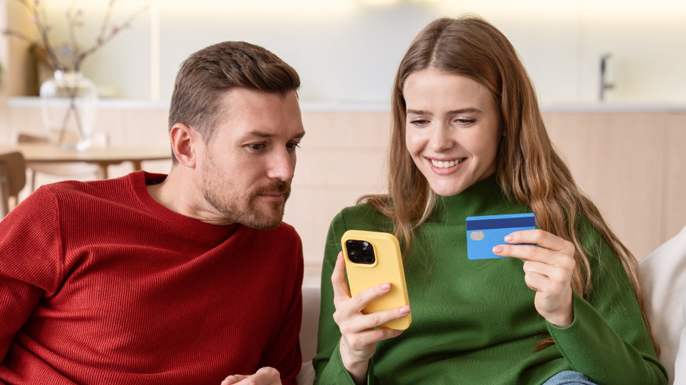 Coppia seduta sul divano guarda lo smartphone mentre la donna tiene in mano una carta di pagamento