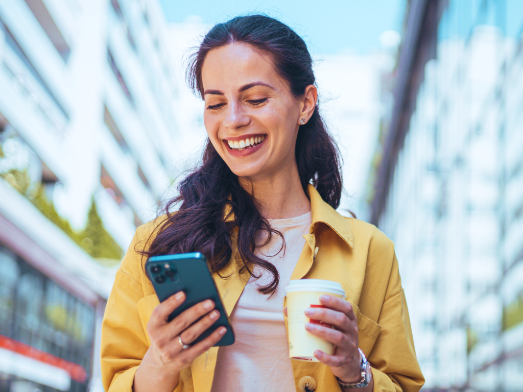 Giovane donna sorridente con giacca gialla guarda lo smartphone tenendo un caffè in mano, in una strada di città