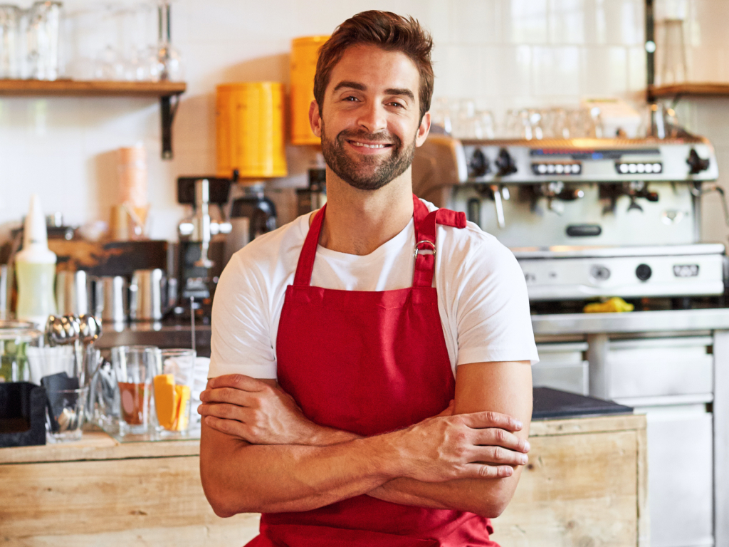 Barista sorridente con grembiule rosso e braccia incrociate dietro il bancone di un bar con macchina del caffè
