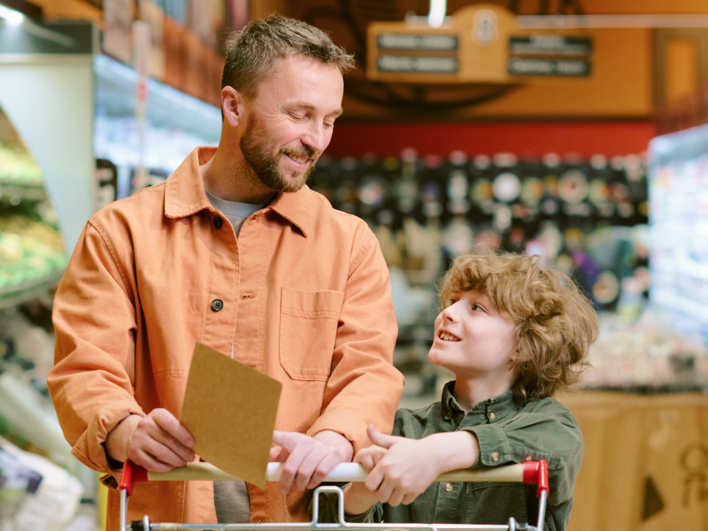 Padre e figlio si sorridono mentre fanno la spesa in un supermercato, con il carrello e la lista in mano