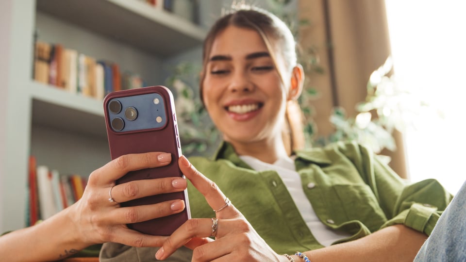 Giovane donna sorridente in camicia verde usa lo smartphone seduta in una stanza luminosa con libreria