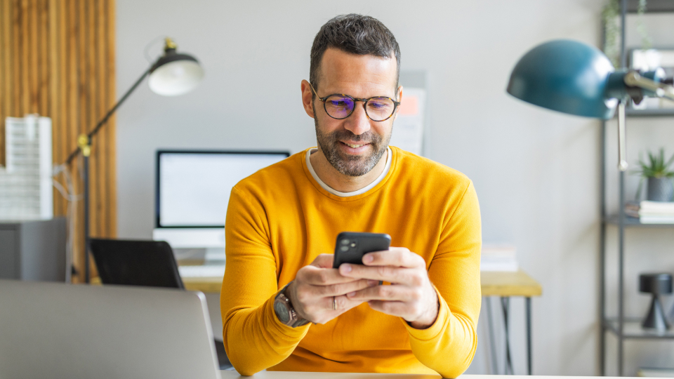 Uomo con occhiali e maglione giallo usa lo smartphone alla scrivania di un ufficio con laptop e monitor