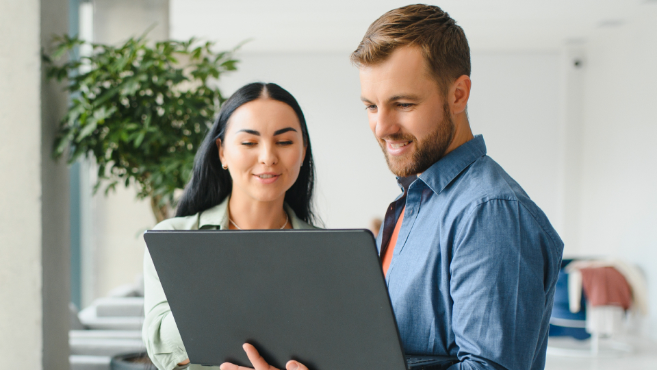 Una donna e un uomo sorridenti guardano insieme lo schermo di un laptop in un ufficio luminoso con piante